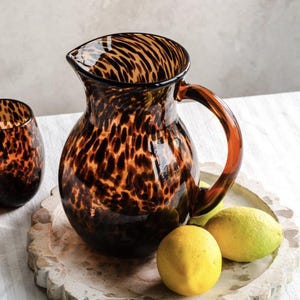 May include: A glass jug and a matching glass tumbler featuring a brown and black tortoiseshell pattern. The jug has a curved handle and a wide spout. Two bright yellow lemons are positioned near the base of the jug.