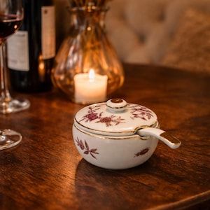 May include: A white ceramic sugar bowl with a lid and a small handle. The bowl is decorated with a floral pattern in shades of purple and gold trim. The bowl is on a dark wooden table.