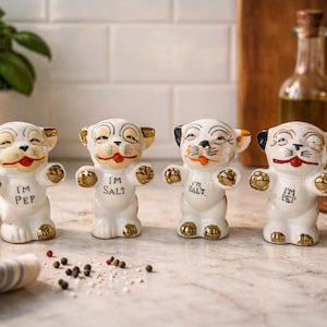 May include: Set of four ceramic salt and pepper shakers shaped like smiling puppies. The white figurines have gold accents on their ears, paws, and feet. Each shaker has a different black ear and the words "I'M PEP" or "I'M SALT" on the front.