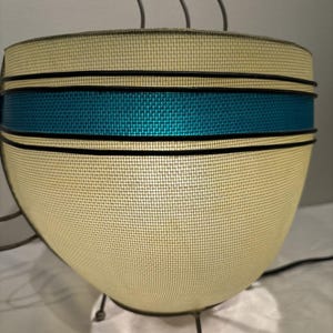 May include: A vintage-style lamp with a cream-colored mesh shade, accented by a teal band and black trim. The lamp has a rounded shape and is supported by three metal legs. The lamp has a retro design.