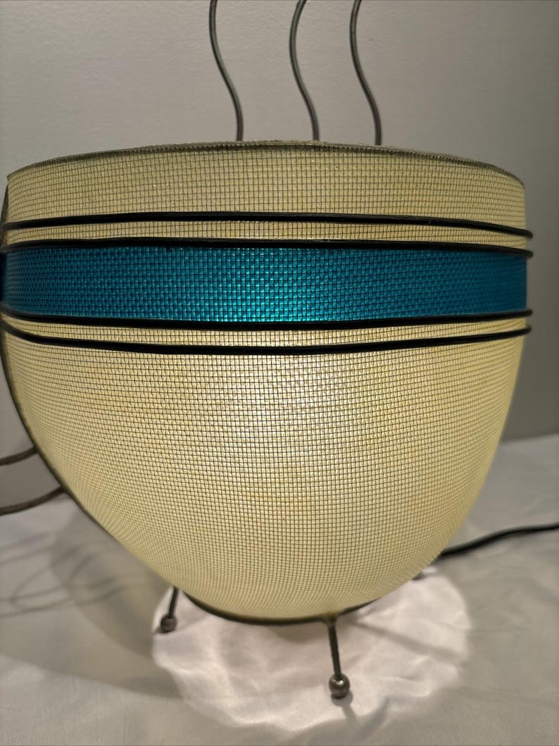 May include: A vintage-style lamp with a cream-colored mesh shade, accented by a teal band and black trim. The lamp has a rounded shape and is supported by three metal legs. The lamp has a retro design.