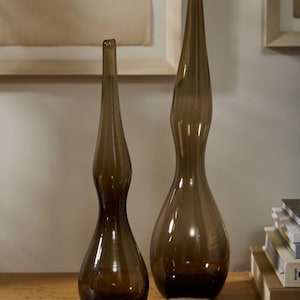 May include: Two brown glass vases with elongated, curved shapes. The vases have a dark brown base that fades to a lighter brown towards the top. They are displayed on a wooden surface, with framed artwork in the background.