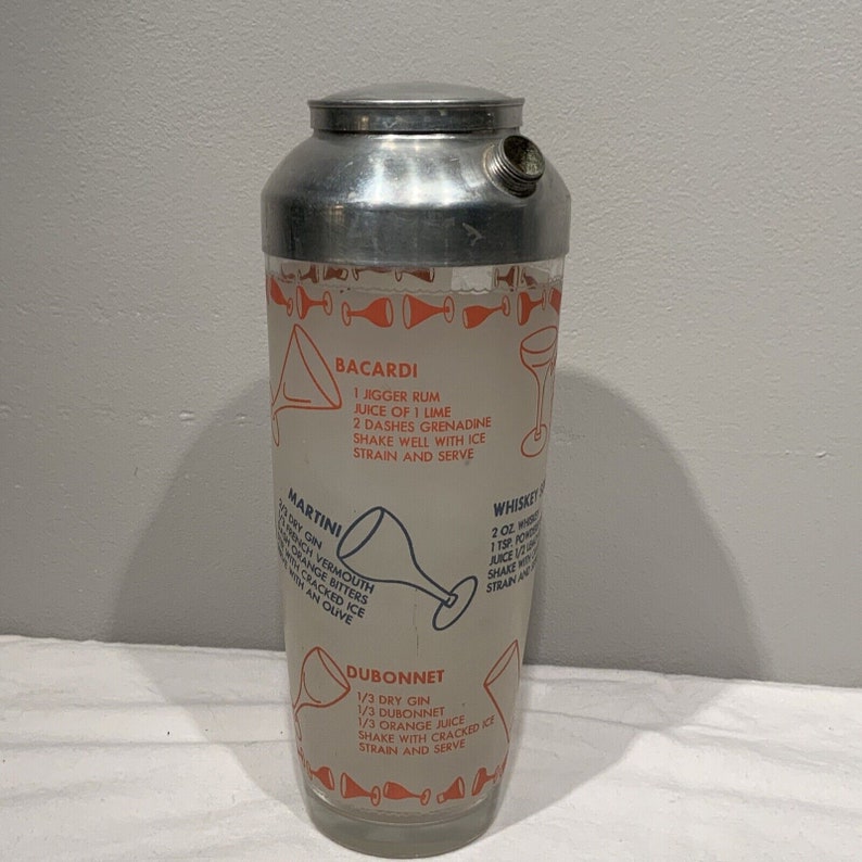 May include: A silver and glass cocktail shaker with a recipe for a Bacardi, Martini, Whiskey, and Dubonnet cocktail printed on the side. The recipes are written in black and orange text.