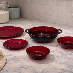 Könnte beinhalten: Eine Sammlung roter Glasservierschalen, darunter Teller und Schalen, die auf einer Marmortheke angeordnet sind. Das Set umfasst einen großen Teller, einen kleinen Teller, eine Schüssel mit Griffen und zwei kleinere Schalen. Ein Glas Mehl befindet sich im Hintergrund.