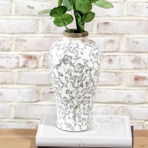 May include: A white ceramic vase with a black and grey floral design, containing green leafy stems. The vase has a brown rim and rests on a white book. The background is a white brick wall.