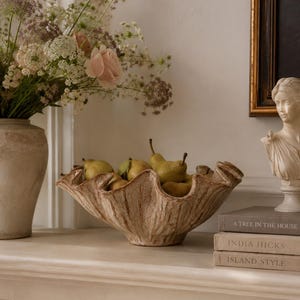 Könnte beinhalten: Eine breite, gewellte Keramikschale mit strukturierter, braun-cremefarbener Oberfläche, gefüllt mit mehreren grünen Birnen. Die Schale steht auf einem Kaminsims neben einer hohen Blumenvase, einer kleinen weißen Büste und drei gestapelten Büchern.