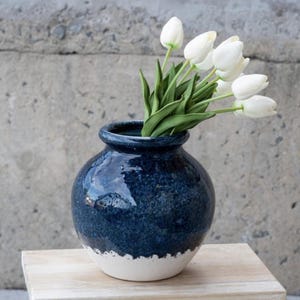 Op de afbeelding: Een ronde, donkerblauwe keramische vaas met een glanzende afwerking, met witte tulpen met groene stelen en bladeren. De bodem van de vaas is wit. De vaas staat op een lichtgekleurd houten oppervlak tegen een grijze betonnen muur.