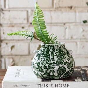 May include: A small, white and green floral patterned vase with two small handles, holding a green fern. The vase sits on a book titled "THIS IS HOME" by Natalie Walton.