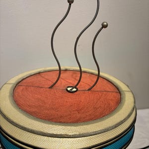 May include: A unique lamp with a cylindrical base and a round top. The top features a rust-colored fabric center, surrounded by a cream-colored border and teal band. Three curved metal rods with ball-shaped tips extend upwards.