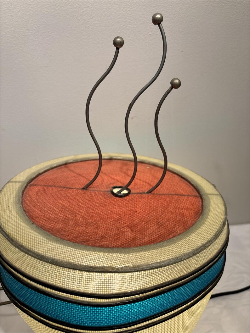 May include: A unique lamp with a cylindrical base and a round top. The top features a rust-colored fabric center, surrounded by a cream-colored border and teal band. Three curved metal rods with ball-shaped tips extend upwards.