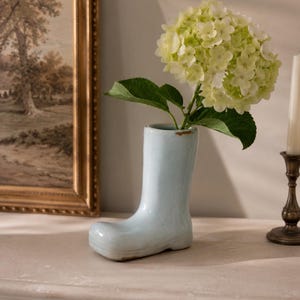 May include: A light blue ceramic boot-shaped vase with a glossy finish and subtle brown distressed detailing along the top rim and toe. The vase holds a single stem of pale green hydrangea flowers with several broad, dark green leaves.
