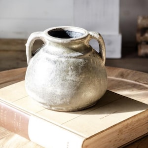 Könnte beinhalten: Eine kleine, runde, cremefarbene Keramikvase mit zwei gebogenen Griffen. Die Vase hat eine strukturierte Oberfläche und ein dunkles Inneres. Sie steht auf einem Vintage-Buch mit hellbraunem Einband.
