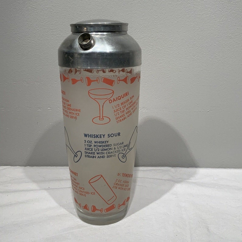 May include: A silver cocktail shaker with a white background and orange text. The shaker has instructions for making a Daiquiri, Whiskey Sour, and a Screwdriver.