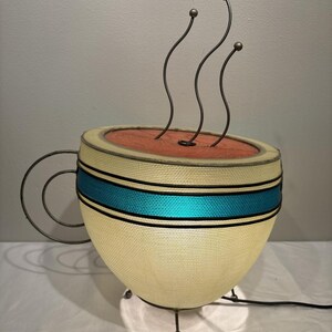 May include: A unique lamp shaped like a coffee cup. The cup is cream-colored with a teal blue stripe and a dark brown rim. Three curved metal rods with ball-shaped tops rise from the top, resembling steam. The lamp has a metal handle and sits on three metal legs.