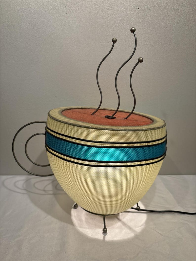 May include: A unique lamp shaped like a coffee cup. The cup is cream-colored with a teal blue stripe and a dark brown rim. Three curved metal rods with ball-shaped tops rise from the top, resembling steam. The lamp has a metal handle and sits on three metal legs.