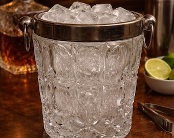 May include: A clear, patterned glass ice bucket filled with ice cubes, featuring a silver rim and handles. The bucket sits on a wooden surface, with a liquor decanter and cocktail shaker in the background.