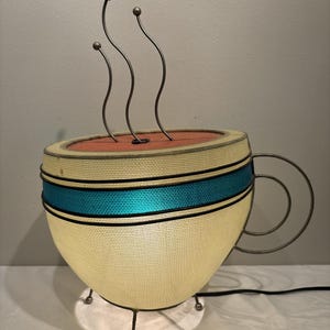 May include: A novelty lamp shaped like a coffee cup. The cup is cream-colored with a teal blue band and a metal handle. Three curved metal rods with ball tips rise from the top, suggesting steam. The lamp sits on a small, white base.