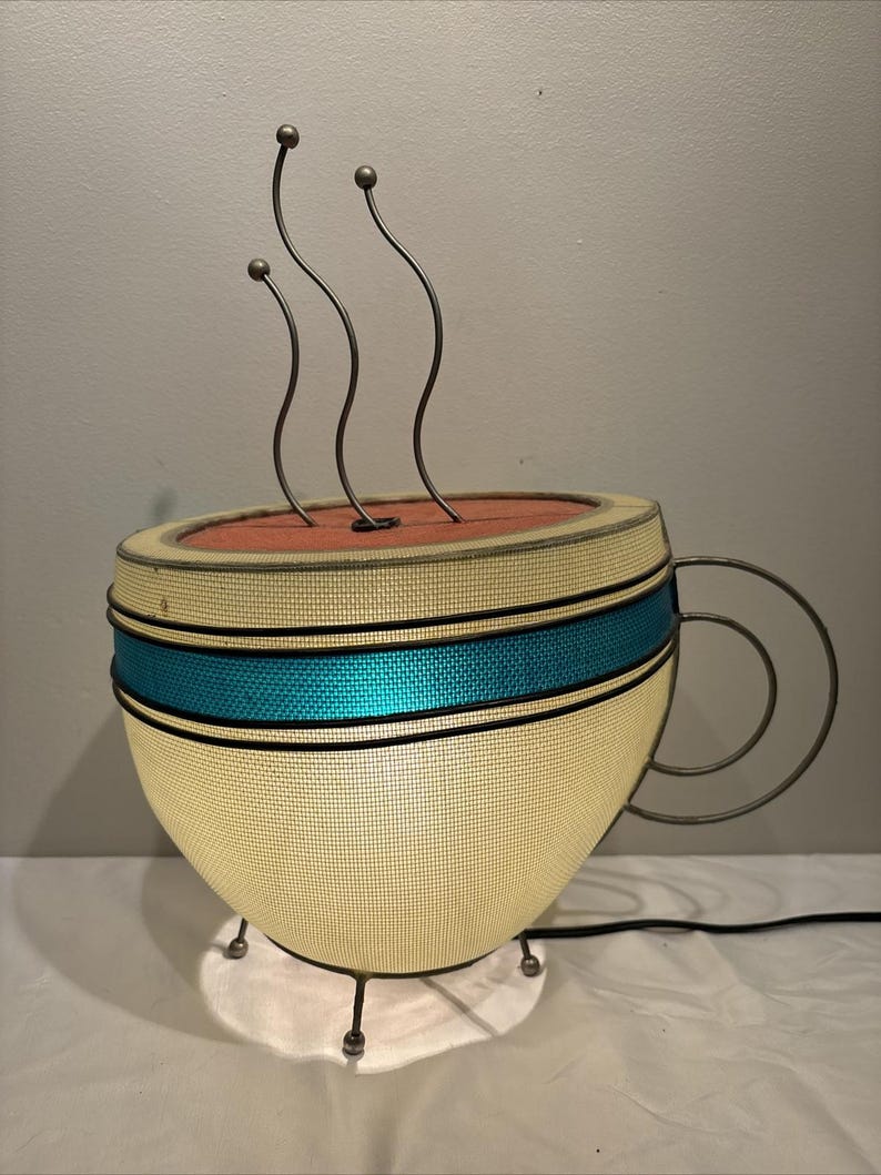 May include: A novelty lamp shaped like a coffee cup. The cup is cream-colored with a teal blue band and a metal handle. Three curved metal rods with ball tips rise from the top, suggesting steam. The lamp sits on a small, white base.