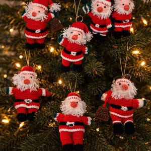 Puede incluir: Adornos navideños de Papá Noel hechos a ganchillo. Cada adorno es rojo con ribetes blancos, un cinturón negro y una barba blanca. Los adornos cuelgan de un árbol de Navidad con luces.