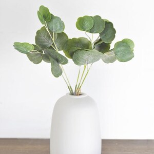 Puede incluir: Un jarrón blanco y redondeado sostiene un ramo de ramas de eucalipto artificiales. Las hojas son de un verde apagado con vetas sutiles, y los tallos son de color verde claro. El jarrón es liso y mate, creando una exhibición simple y elegante.