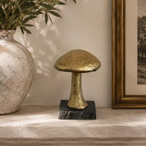 Pode incluir: Uma escultura decorativa de cogumelo com um chapéu e caule dourados texturizados, montada sobre uma base quadrada de pedra cinzenta escura com veios. O objeto tem um acabamento metálico com estrias verticais visíveis no caule.