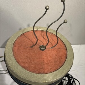 May include: A vintage, round, decorative object with a rust-colored center and a light green border. Three curved metal stems with silver ball tips extend upwards. The object has a blue stripe and a black cord.