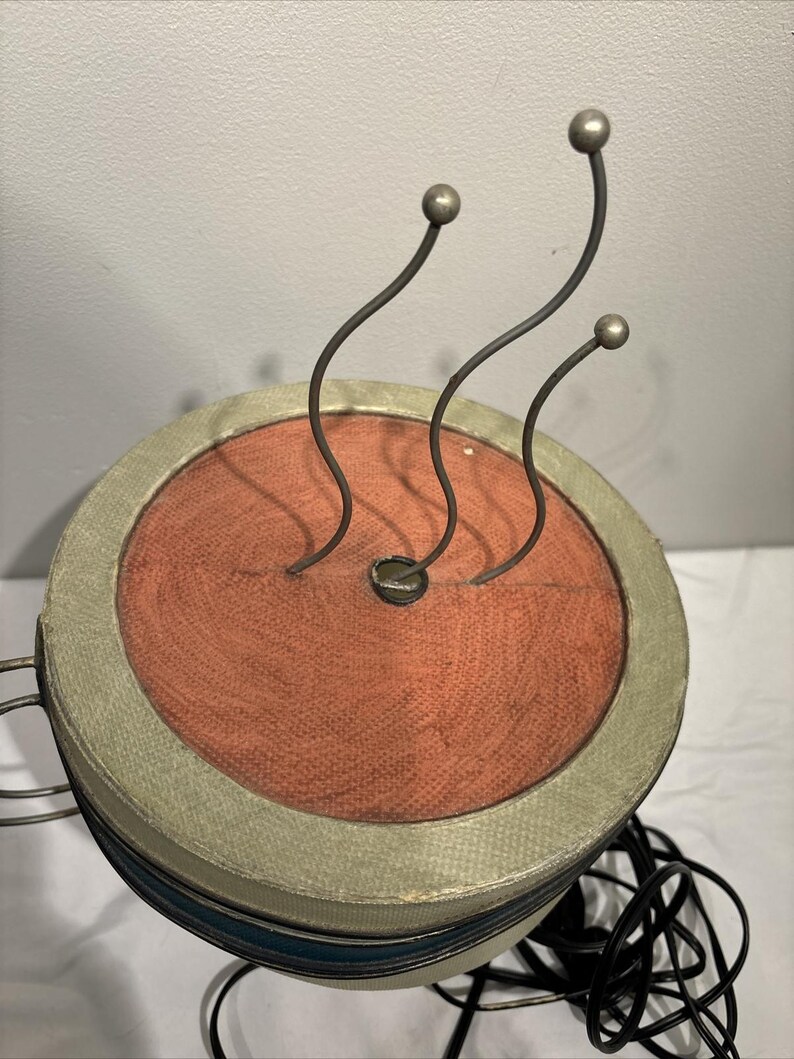 May include: A vintage, round, decorative object with a rust-colored center and a light green border. Three curved metal stems with silver ball tips extend upwards. The object has a blue stripe and a black cord.