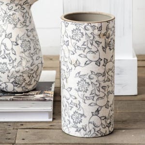 May include: A cylindrical white vase with a floral pattern in gray. The vase has a slightly textured surface and a neutral-colored rim. The design features stylized flowers and leaves.