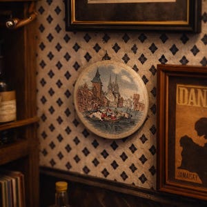 Puede incluir: Una obra de arte redonda enmarcada que representa un paisaje urbano europeo con un río y barcos. La obra está montada en una pared estampada, con otras piezas enmarcadas y una botella de licor visible en el fondo. La paleta de colores es apagada, con tonos cálidos.