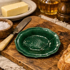 Peut inclure: Un plat à beurre vert en céramique en forme de poisson sur une planche à découper en bois. Le plat présente un motif de poisson détaillé et un bord festonné. Autres articles : beurre, sel, couteau et pot de miel.