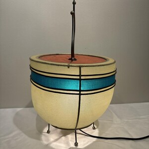 May include: A mid-century modern table lamp with a cream-colored mesh shade, accented by a turquoise band and black trim. The lamp has a red interior and a curved metal stem with three spherical accents. It rests on three small metal legs.