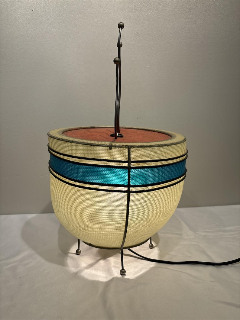 May include: A mid-century modern table lamp with a cream-colored mesh shade, accented by a turquoise band and black trim. The lamp has a red interior and a curved metal stem with three spherical accents. It rests on three small metal legs.