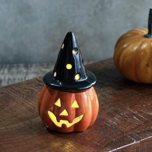 Pode incluir: Decoração de Halloween em cerâmica. Apresenta uma abóbora laranja com um rosto esculpido e um chapéu de bruxa preto com recortes de pontos amarelos. A abóbora está sobre uma superfície de madeira, com outra abóbora ao fundo.