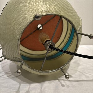 May include: A vintage, spherical lampshade with a textured, light beige exterior. The interior features a colorful design with orange, teal, and cream stripes. The metal frame has small, silver ball accents. A black power cord extends from the lamp.