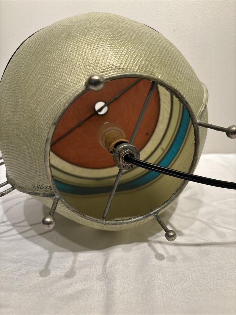 May include: A vintage, spherical lampshade with a textured, light beige exterior. The interior features a colorful design with orange, teal, and cream stripes. The metal frame has small, silver ball accents. A black power cord extends from the lamp.