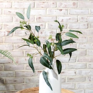 May include: A white ceramic vase holds a branch of artificial eucalyptus with dark green, elongated leaves and small clusters of light green buds. The stems are thin and brown, branching out to support the foliage in a natural arrangement.