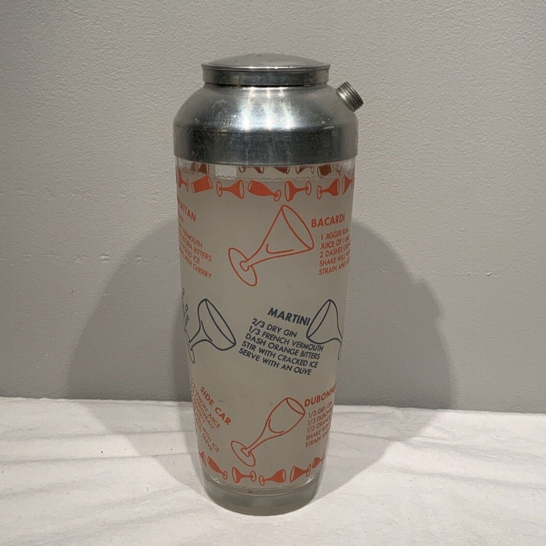 May include: A vintage silver cocktail shaker with a glass body and a printed recipe for a Martini, Bacardi, Side Car, and Dubonnet. The recipes are printed in orange and black text on a white background.