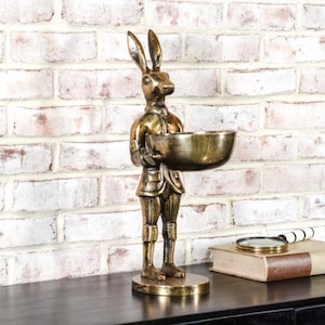 Puede incluir: Una figura dorada de un conejo sosteniendo un cuenco. El conejo viste un atuendo de estilo vintage y se apoya sobre una base redonda. La figura se encuentra frente a una pared de ladrillos blancos.
