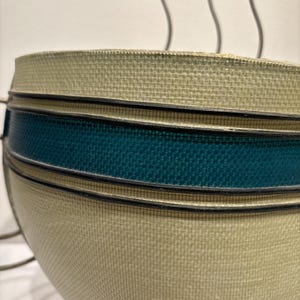 May include: Close-up of a beige and teal woven bowl. The bowl has a rounded shape with a textured surface. A teal band is accented with black trim. The bowl is likely a decorative item or a serving piece.