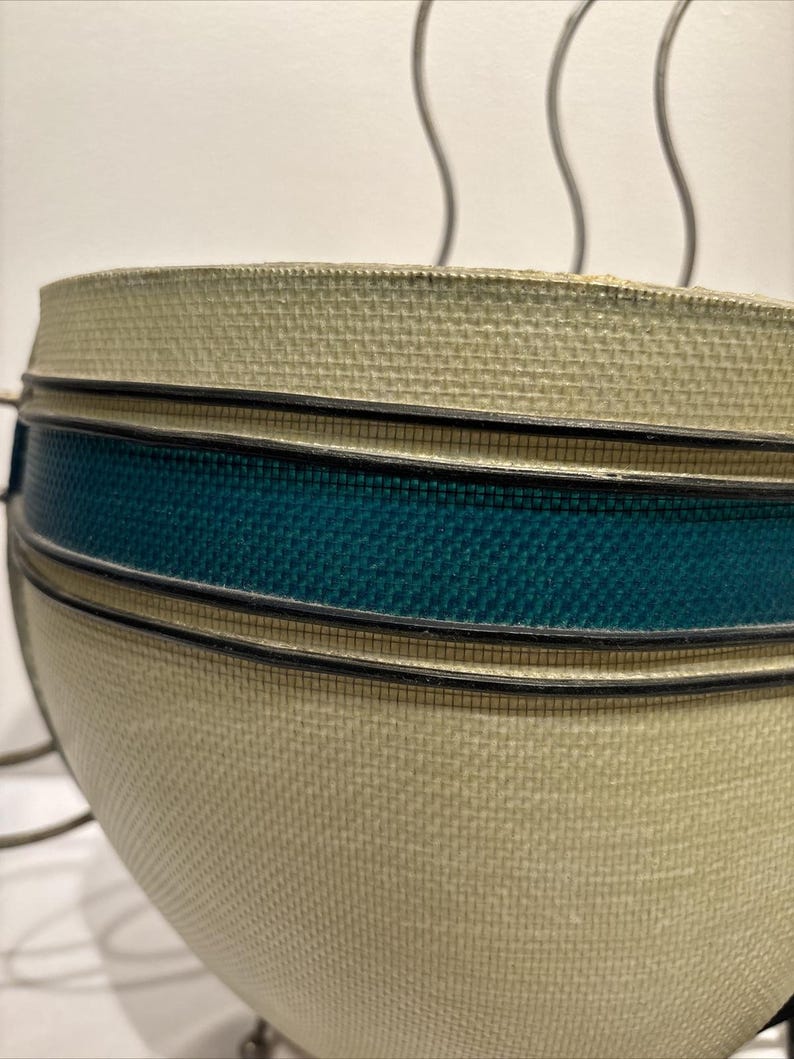 May include: Close-up of a beige and teal woven bowl. The bowl has a rounded shape with a textured surface. A teal band is accented with black trim. The bowl is likely a decorative item or a serving piece.