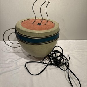 May include: A decorative coffee cup sculpture with a beige and teal exterior, featuring a red interior and three metal steam accents. The cup has a metal handle and a black power cord.