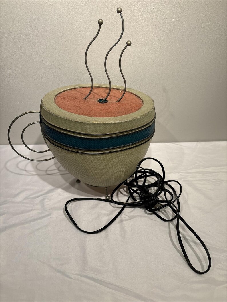 May include: A decorative coffee cup sculpture with a beige and teal exterior, featuring a red interior and three metal steam accents. The cup has a metal handle and a black power cord.