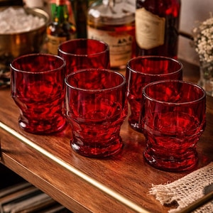 May include: Six red, textured drinking glasses on a wooden serving tray. The glasses are cylindrical with a textured base. Bottles of liquor and a bowl of ice are in the background, suggesting a bar or cocktail setting.