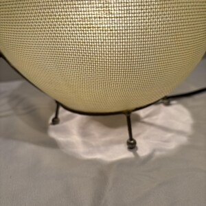 May include: A mid-century modern style lamp with a spherical, off-white mesh shade. The lamp has a dark metal frame and three small legs. The light shines through the mesh, creating a soft glow. The lamp is a decorative lighting fixture.