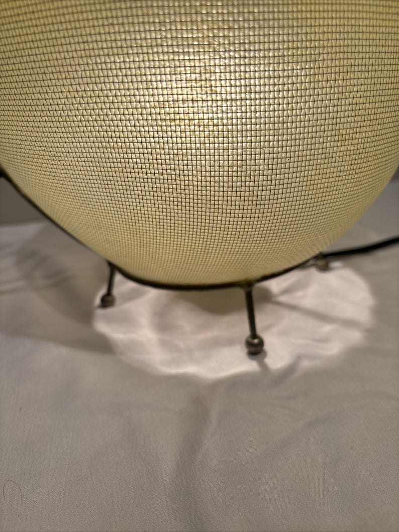 May include: A mid-century modern style lamp with a spherical, off-white mesh shade. The lamp has a dark metal frame and three small legs. The light shines through the mesh, creating a soft glow. The lamp is a decorative lighting fixture.