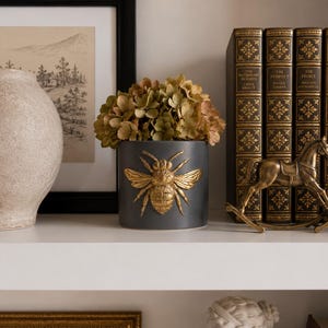 Può includere: Un vaso in ceramica grigio scuro con un design a forma di ape dorata, pieno di fiori secchi. Il vaso è su una mensola bianca, accanto a opere d'arte incorniciate e libri decorativi. È presente anche una statuina di cavallo a dondolo dorato.