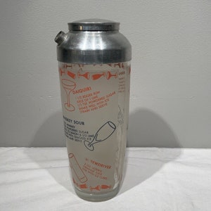 May include: A silver cocktail shaker with a glass insert. The glass insert has a white background with orange and blue text listing cocktail recipes, including Daiquiri, Whiskey Sour, and Screwdriver.