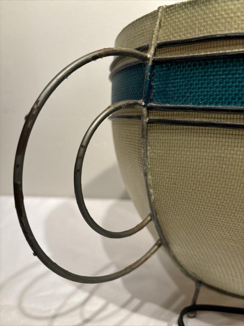 May include: A vintage-style decorative bowl with a woven beige exterior and a teal band. The bowl features a metal frame and handle, adding a touch of retro charm. The design is reminiscent of mid-century modern decor.