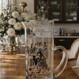 May include: A clear glass pitcher with a curved handle, featuring a black illustration of tennis players. The pitcher has handwritten text at the top. The pitcher is sitting on a wooden table.