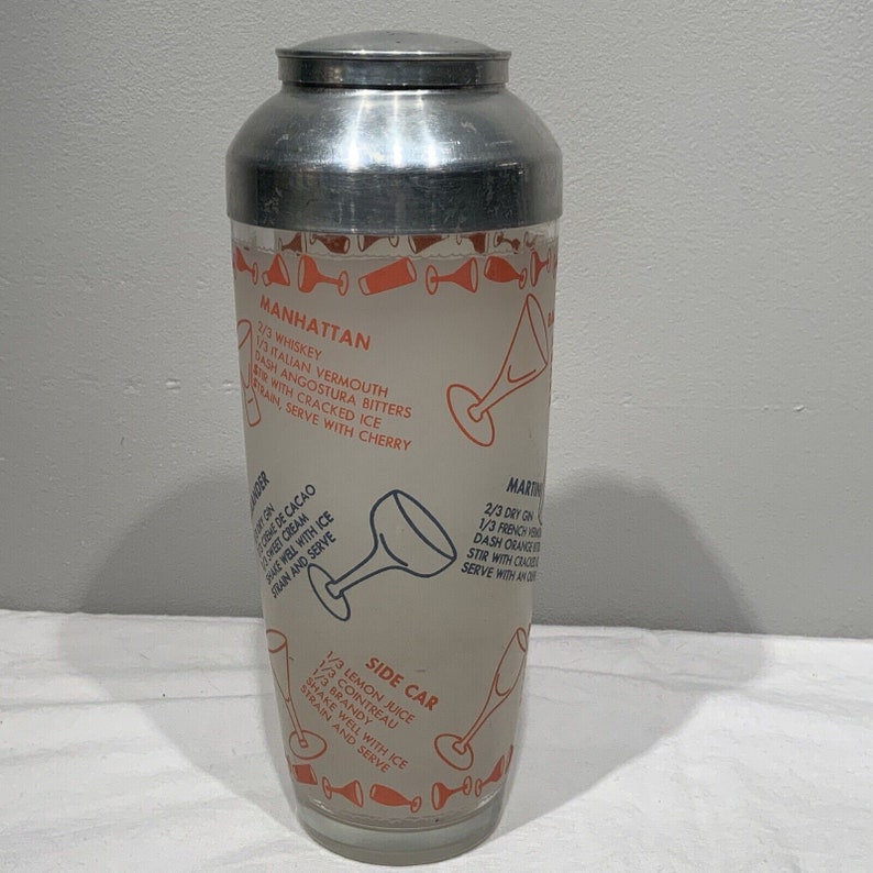 May include: A silver cocktail shaker with a glass insert featuring cocktail recipes printed in orange and blue. The recipes include Manhattan, Martini, and Side Car.